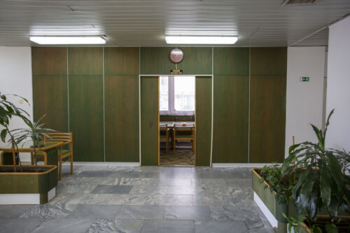 The original green-stained wall panelling, planters, and furniture complete the entrance foreground of the meeting hall