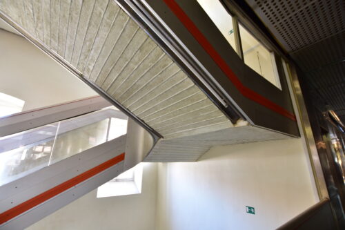 Železobetónové ramená schodiska dotvára atypicky modelované zábradlie, akcentujúce červené línie a dizajnovo nadčasové lineárne svietidlá