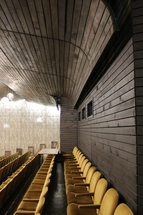 Výtvarne nadčasový, lineárne členený drevený podhľad a obklad v kombinácii s plasticky štruktúrovaným stenovým obkladom optimalizujú priestorovú akustiku kino-divadelnej sály